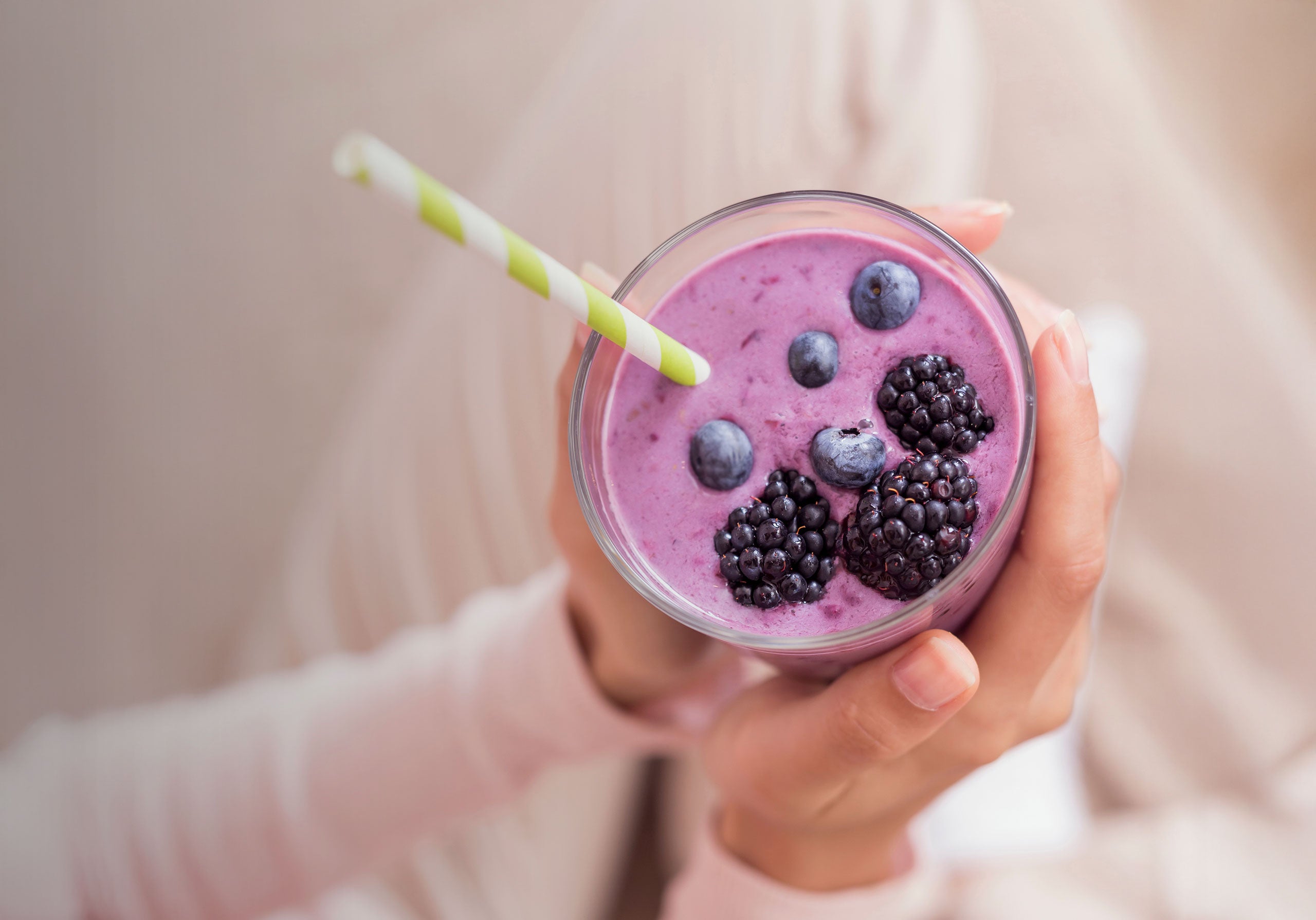 Hand holding a purple smoothie with berries and a straw against a neutral background